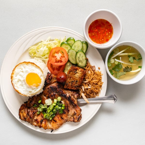 Vietnamese grilled pork chop with fried egg, rice, vegetables, and dipping sauces on a white plate.