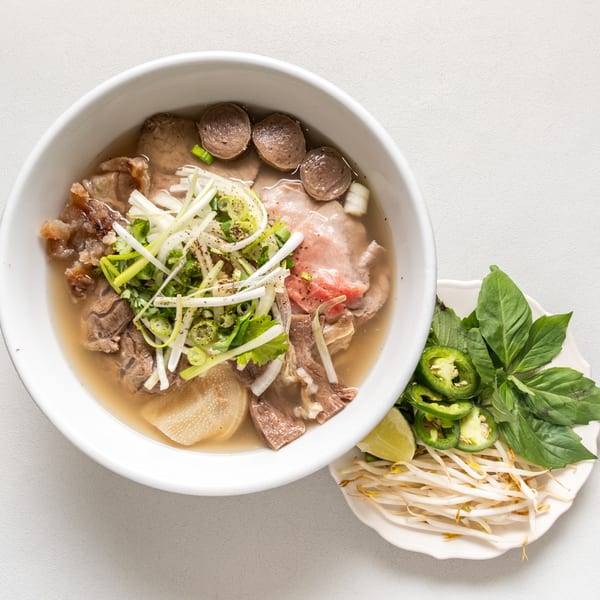 A bowl of Pho Vietnam with beef, meatballs, and fresh herbs, served with a side of garnishes.