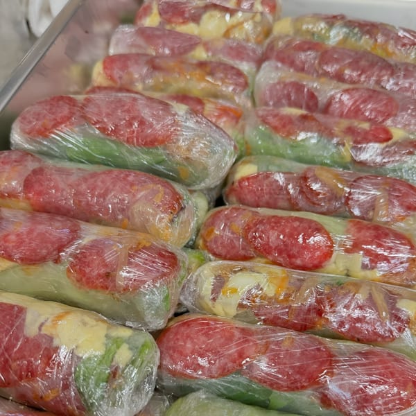 Rows of Vietnamese spring rolls with sausage and vegetables, wrapped in plastic, in a metal tray.