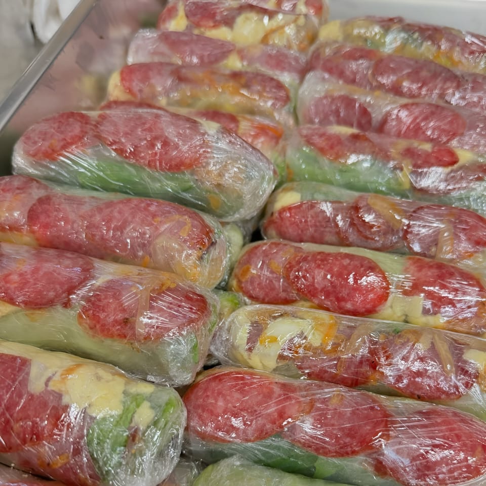 Rows of Vietnamese spring rolls with sausage and vegetables, wrapped in plastic, in a metal tray.